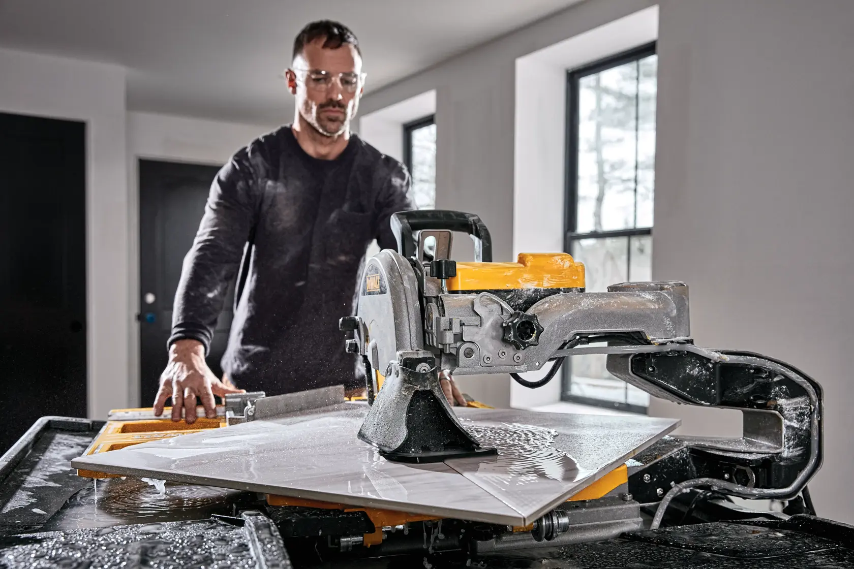 A person using a DEWALT D36000 tile saw to cut a large tile on a worktable in a room with two windows.