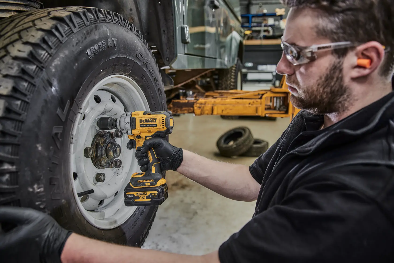 Eine Person mit Handschuhen benutzt einen kabellosen DEWALT Schlagschrauber (Modell DCF891P2G), um an den Radmuttern eines Fahrzeugreifens in einer Werkstatt zu arbeiten. Das Gesicht der Person ist aus Datenschutzgründen unscharf.