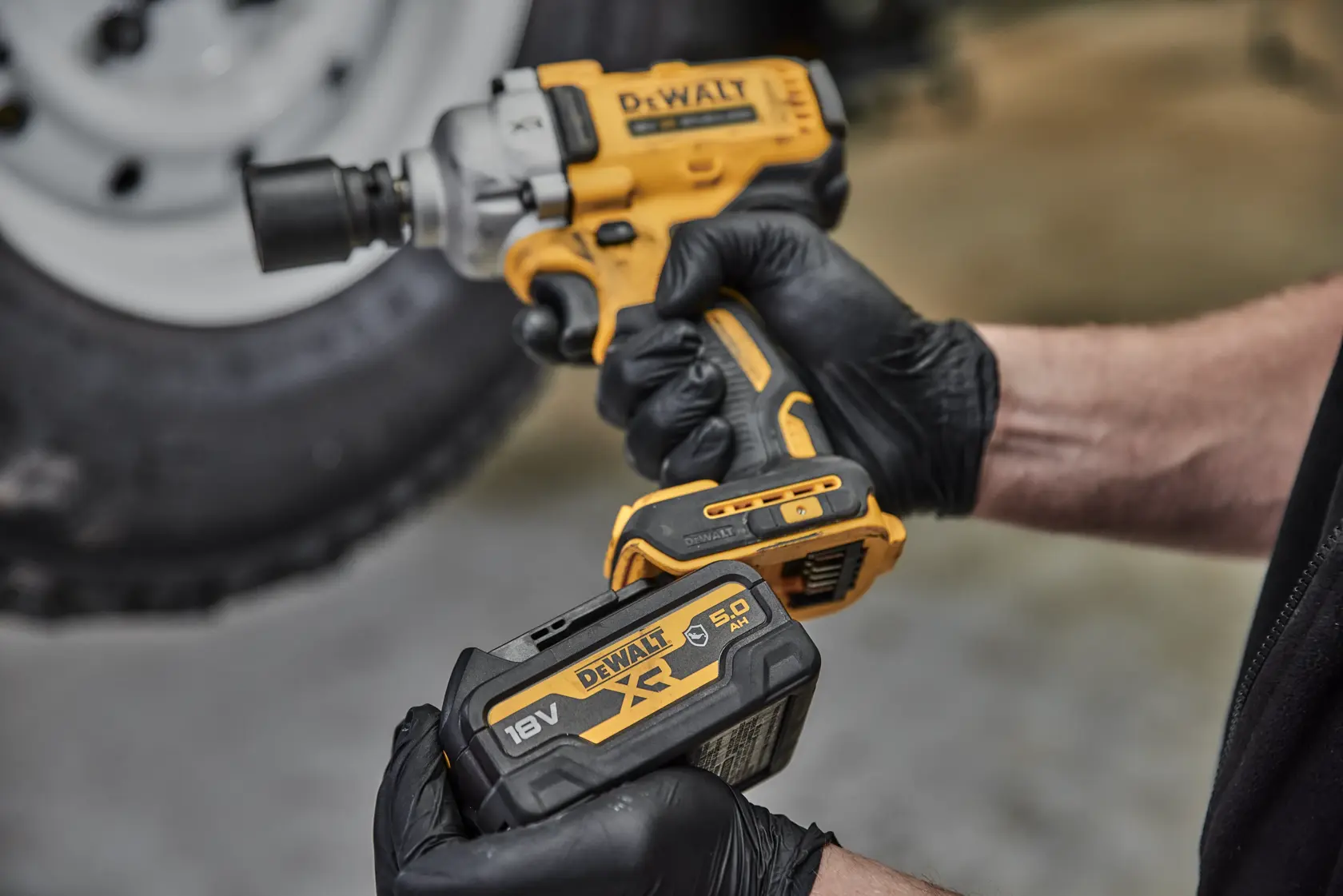 Eine Person mit schwarzen Handschuhen hält einen gelb-schwarzen DEWALT Akku-Schlagschrauber und einen DEWALT XR 18V 5,0 Ah Lithium-Ionen-Akku. Im Hintergrund ist ein verschwommenes Reifenrad zu sehen.