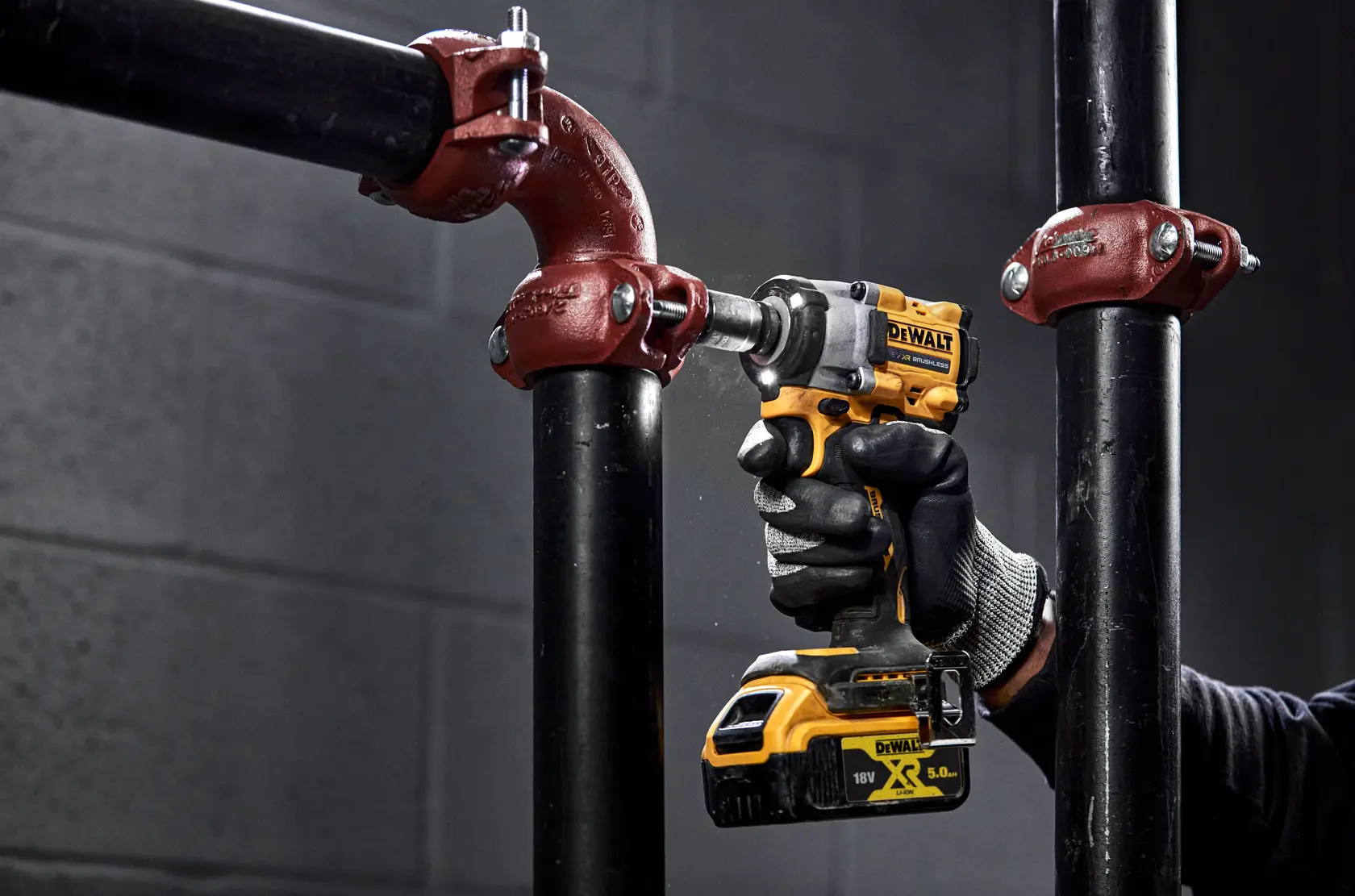 Una persona con guantes negros utiliza una llave de impacto inalámbrica DEWALT amarilla y negra para apretar un perno en un accesorio de tubería metálica en un entorno industrial.