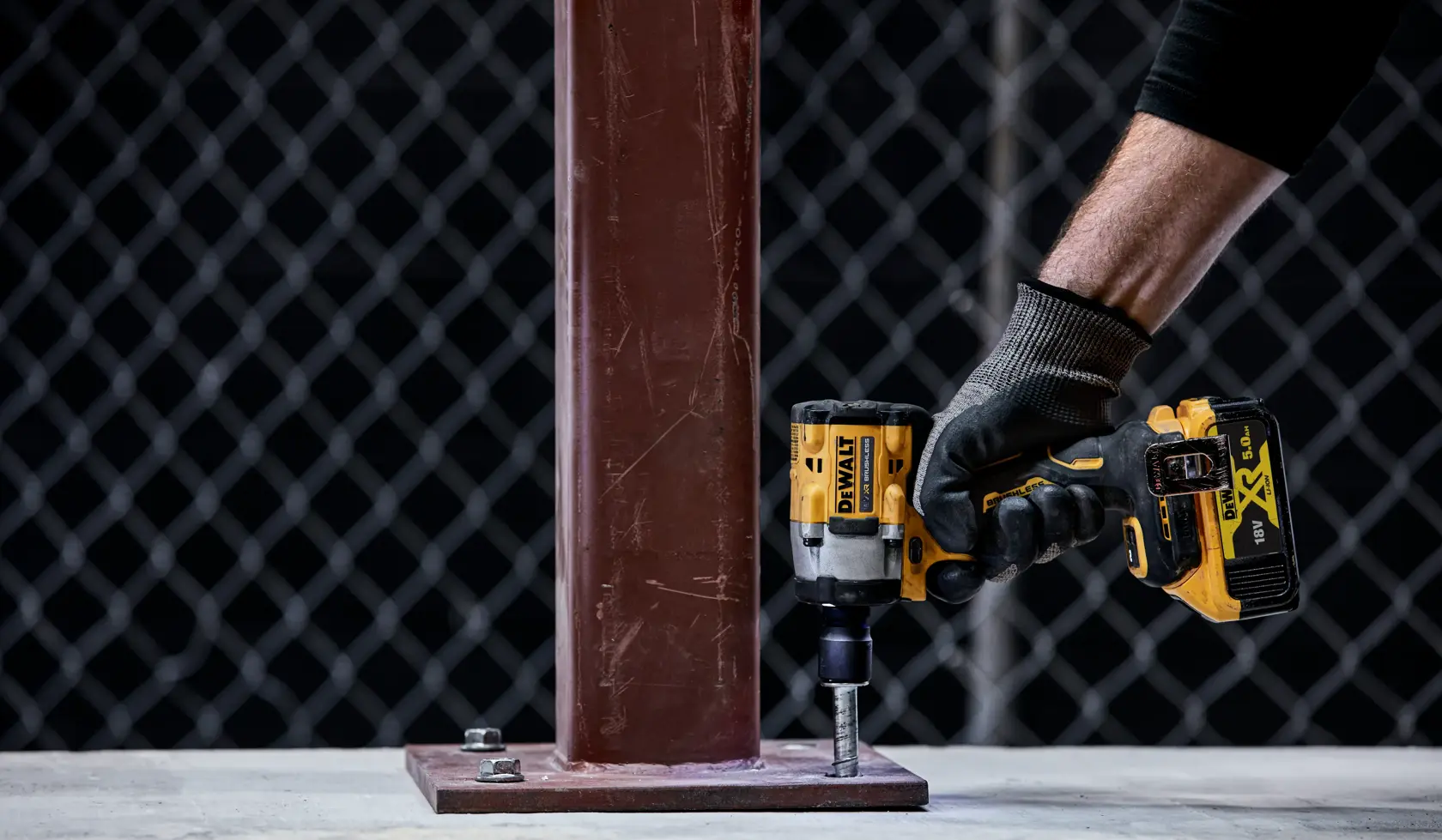Una mano enguantada utiliza una llave de impacto inalámbrica DEWALT (modelo DCF921P2T) para apretar un perno en la base de una columna metálica en un entorno industrial.