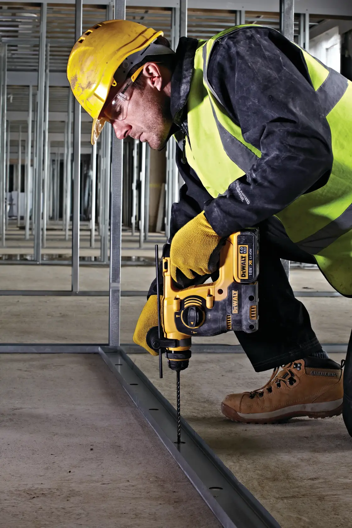 Un trabajador de la construcción con casco amarillo, chaleco de seguridad, guantes y botas está utilizando un martillo rotativo inalámbrico DEWALT DCH253 para perforar un marco metálico en una obra.