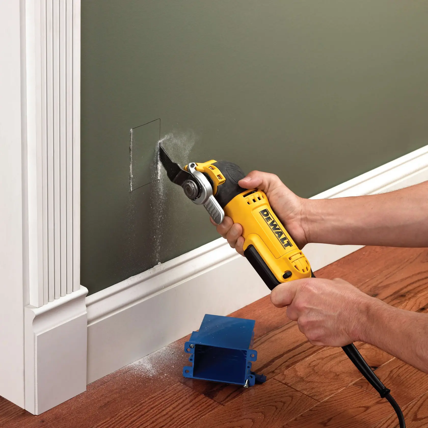 Herramienta DEWALT cortando un agujero en pared, sostenida por manos. Caja azul en el suelo. Polvo visible.