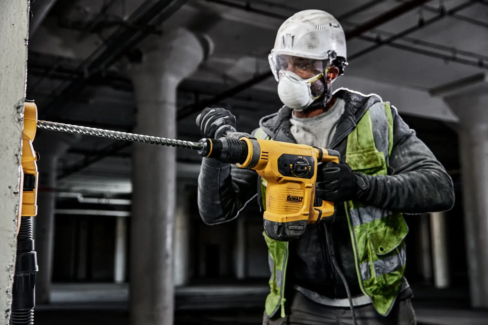 A construction worker wearing protective gear is using a yellow DEWALT hammer drill to drill into a concrete pillar on a construction site.