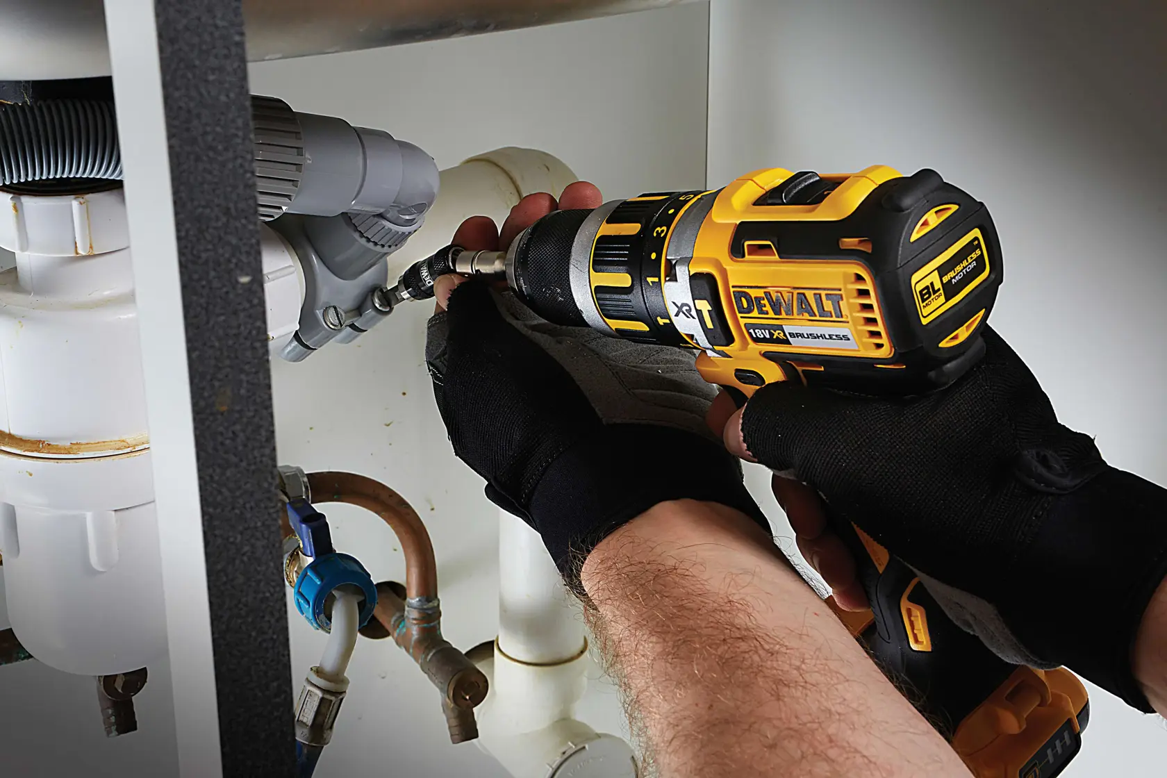 Person uses DEWALT drill to work on pipes under a sink, wearing black gloves for safety.