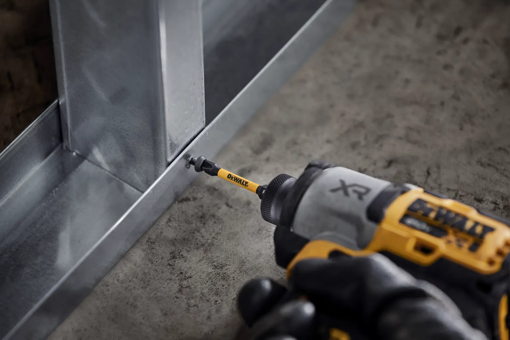 DEWALT drill driving a screw into a metal frame on a concrete floor using a yellow bit.