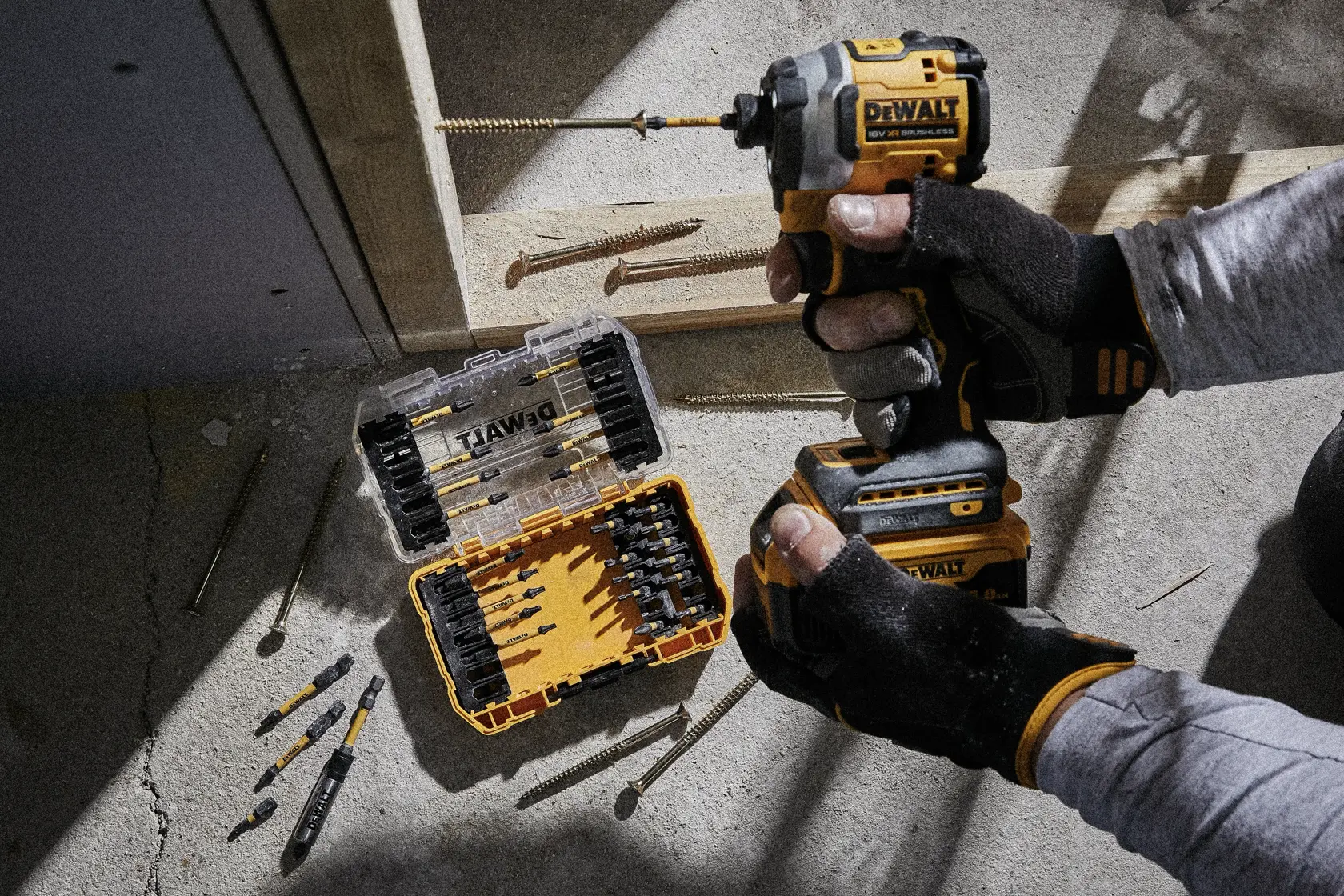 Person arbeitet mit DEWALT-Bohrer, offene Bitbox und Schrauben auf Betonboden einer Baustelle.