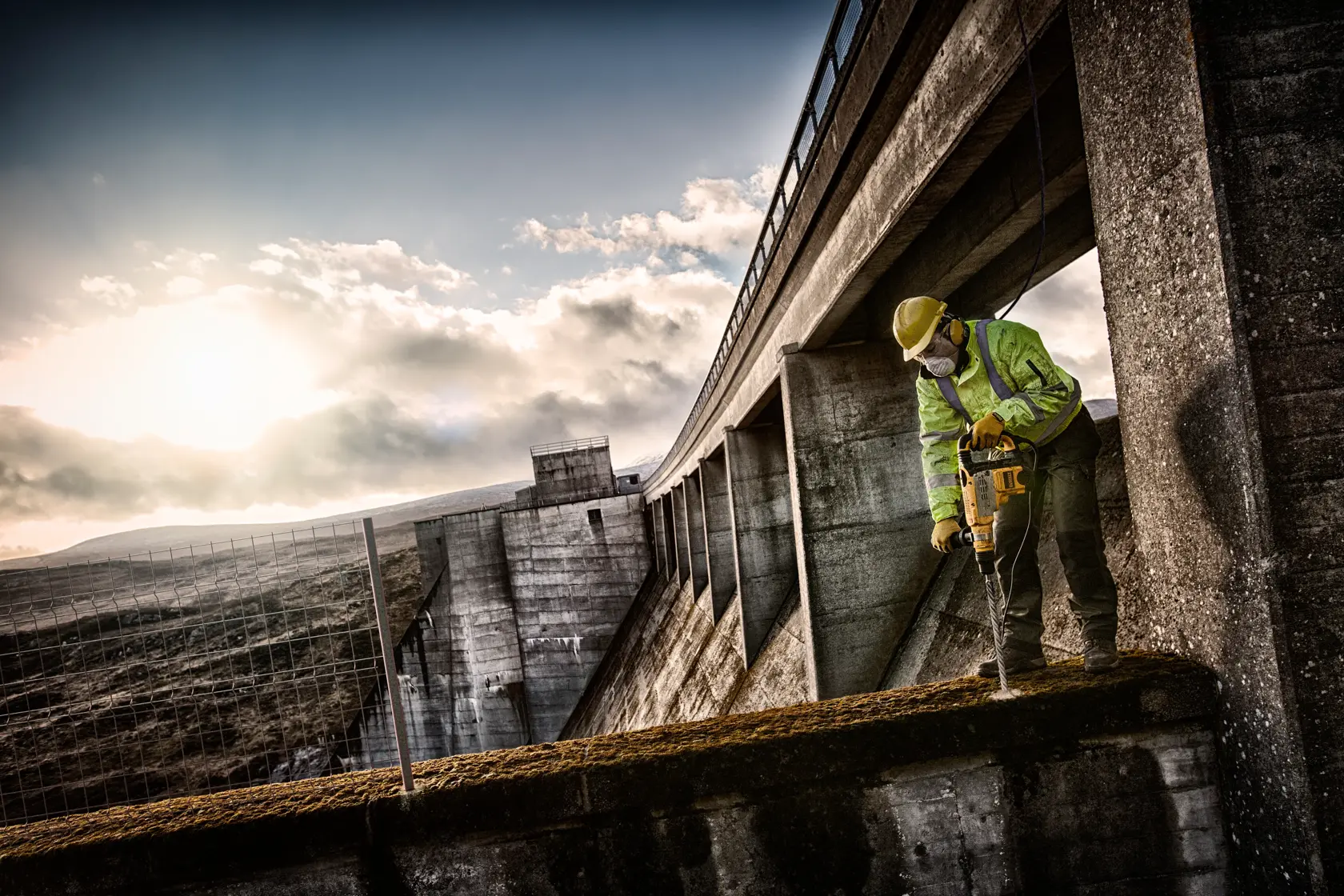 Arbeiter nutzt DEWALT-Bohrer draußen auf Betonstruktur, mit Schutzkleidung unter teils bewölktem Himmel.