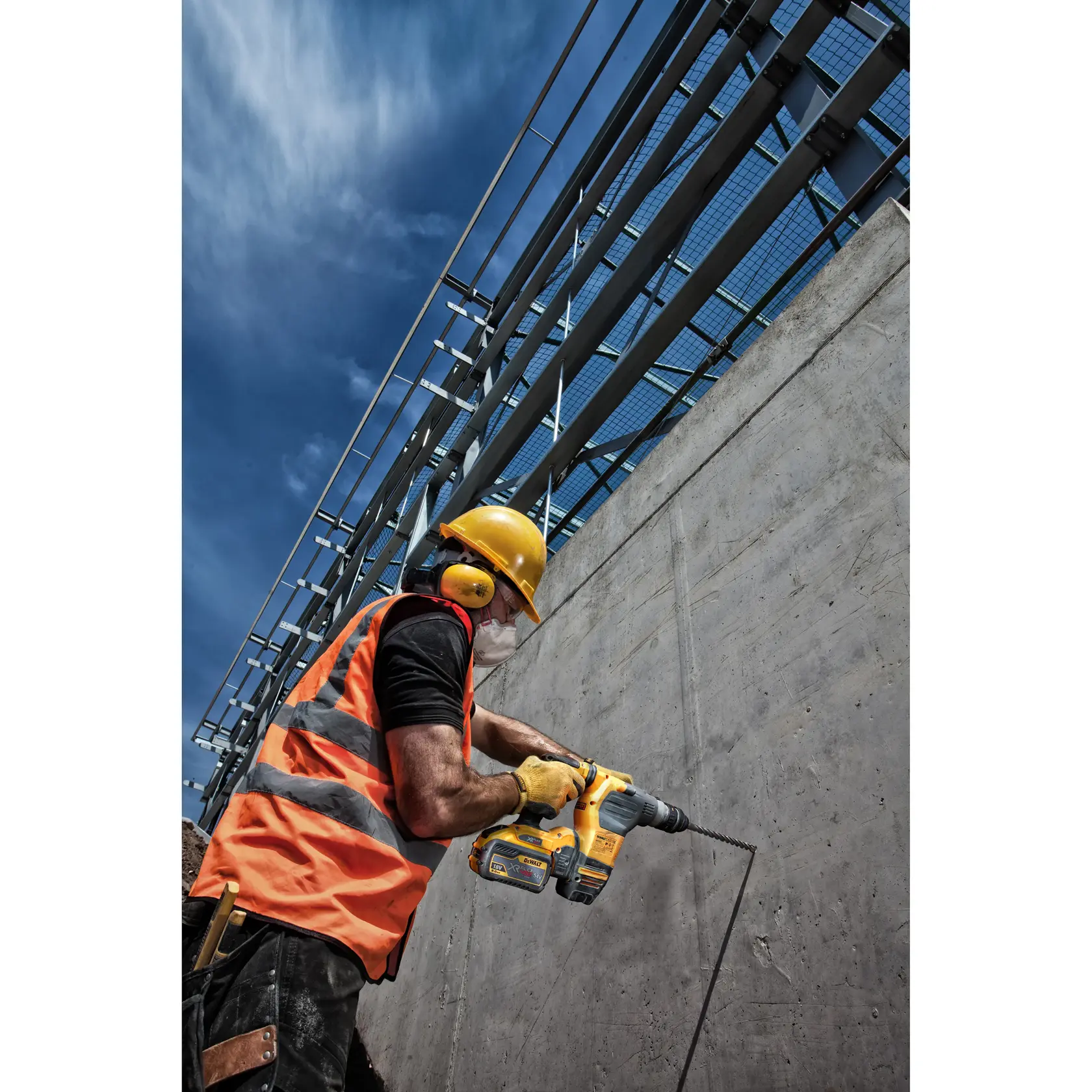Trabajador con equipo de seguridad usa taladro DEWALT en pared de concreto bajo cielo despejado.