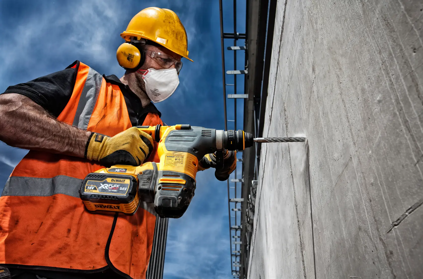 Trabajador perfora concreto con taladro inalámbrico DEWALT. Lleva chaleco naranja y equipo de seguridad.