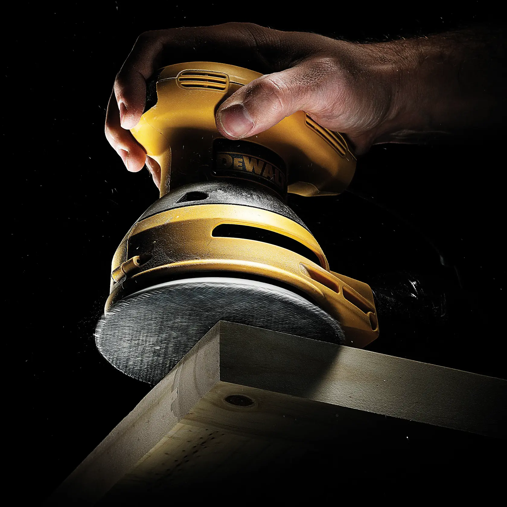 Hand using a DEWALT sander to smooth the edge of a piece of wood, close-up under dramatic lighting.