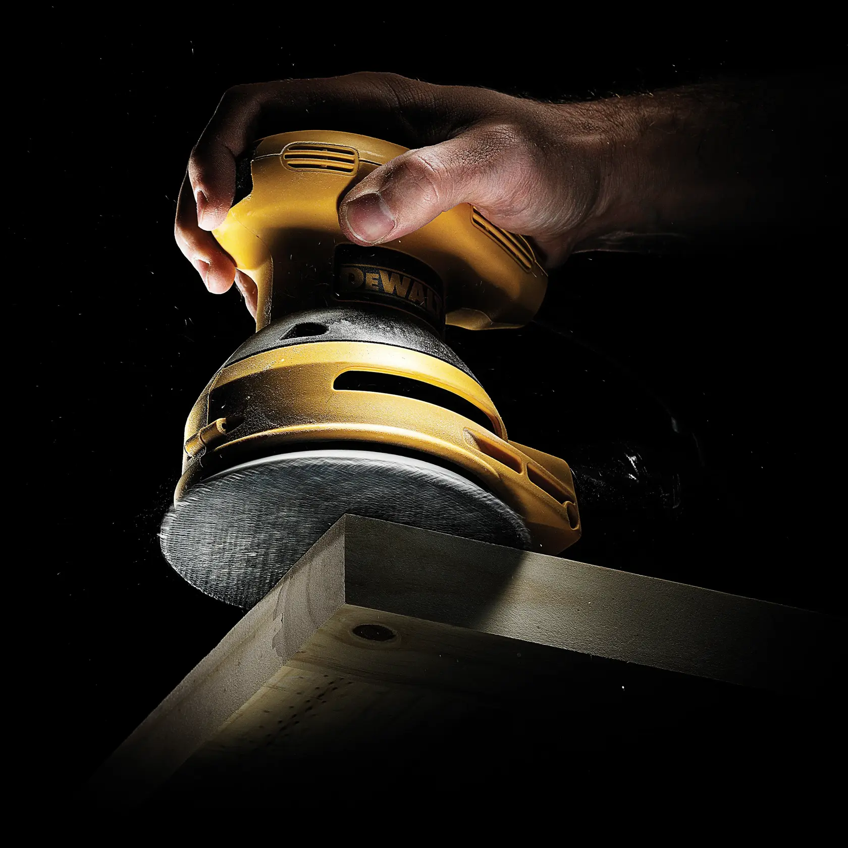 A DEWALT hand sander sanding a wooden plank, held firmly in a hand against a dark background.