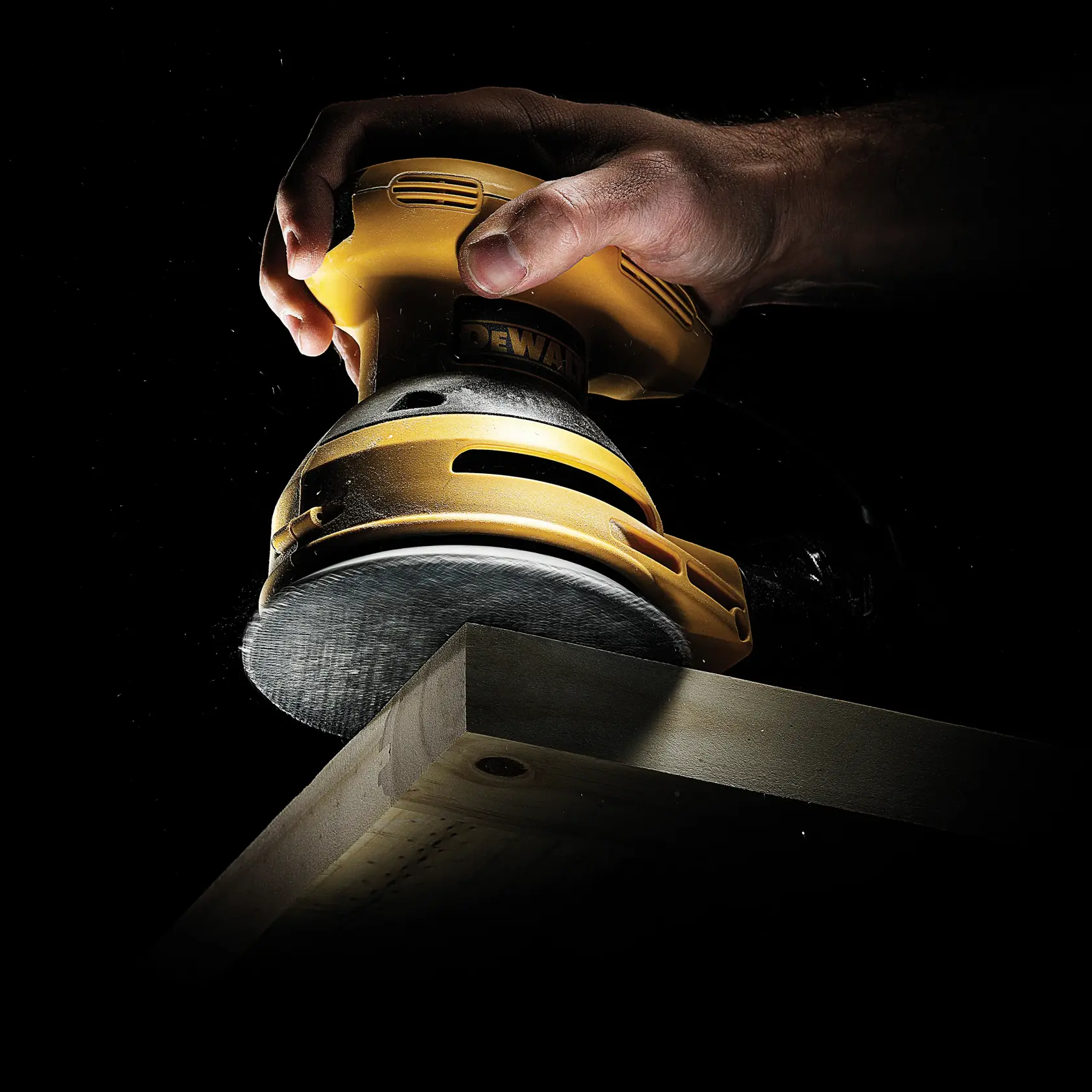 A DEWALT sander is used to smooth a wooden board edge, held by a hand against a dark background.